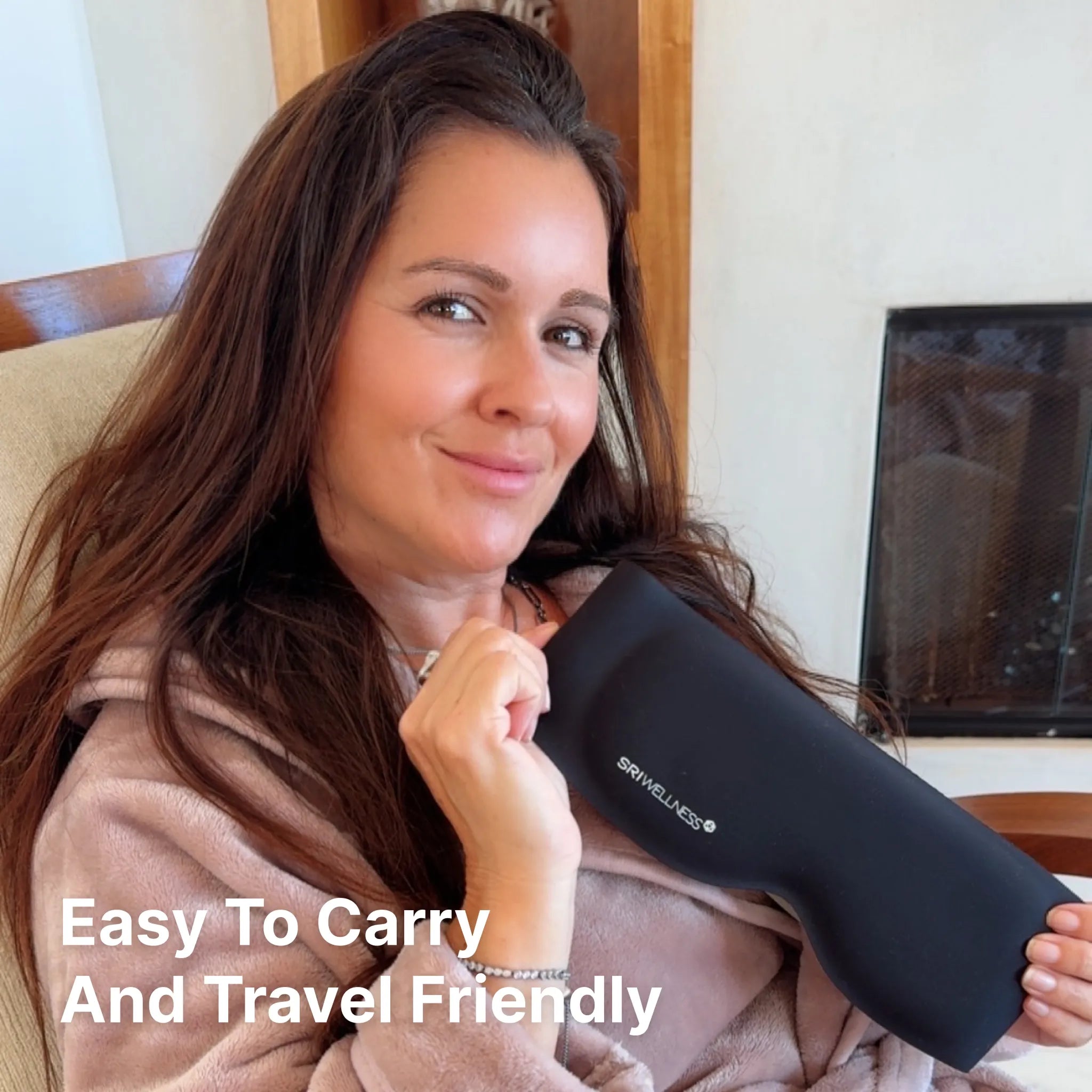 A woman holding a black travel neck pillow with the brand name 'SRI Wellness' visible on the side. She is wearing a pinkish-brown robe in an indoor setting, with text overlay that reads 'Easy To Carry And Travel Friendly'.