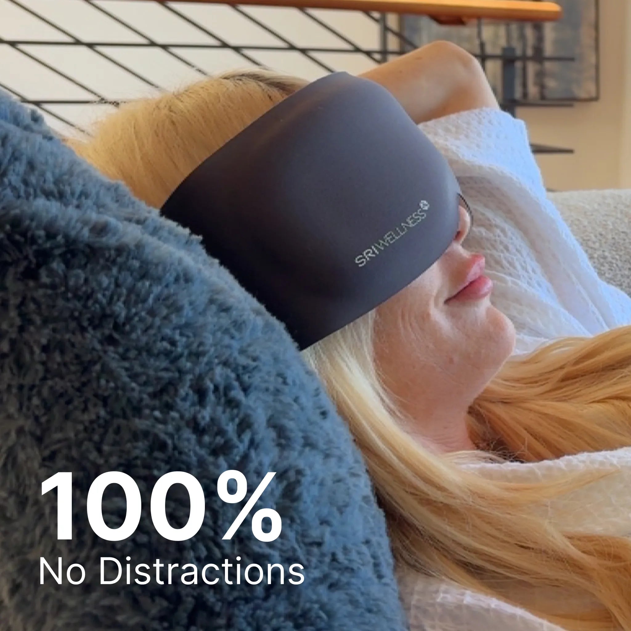 A woman reclining with her eyes closed, wearing a dark gray SRI Wellness sleep mask. She is resting on a blue textured blanket with a white towel, in a cozy indoor setting.
